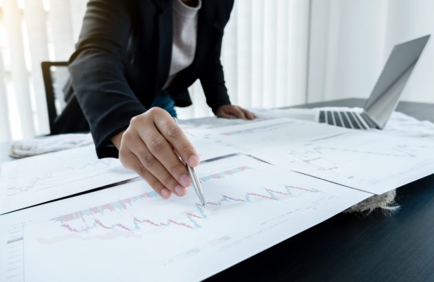 Profissional com caneta analisa gráficos em documentos sobre mesa. Laptop ao fundo.