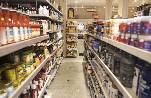 Corredor de mercado com prateleiras repletas de produtos e um expositor de pães no centro.