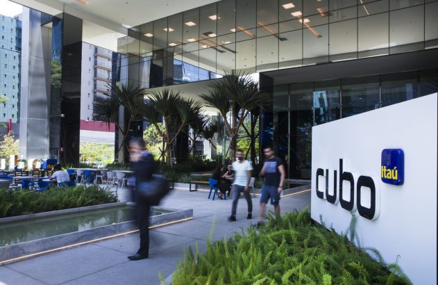 Pessoas caminham e interagem em área externa de prédio moderno com fachada de vidro, palmeiras e um letreiro "cubo Itaú".