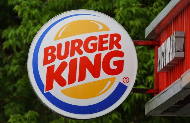 Sinal circular do Burger King com logotipo de sanduíche, contra folhagem verde e parte de prédio vermelho.