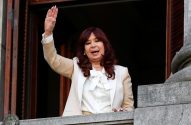 Cristina Kirchner acena de sacada de pedra, sorrindo. Veste blazer claro e blusa branca com laço.