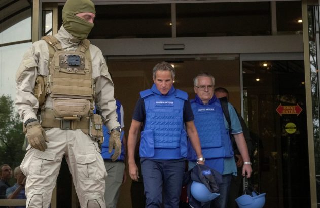 Homens da ONU com coletes azuis saem de prédio, escoltados por segurança de balaclava e uniforme tático.