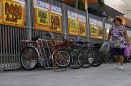 Mulher com sacolas anda por cartazes de ofertas e bicicletas encostadas numa grade.
