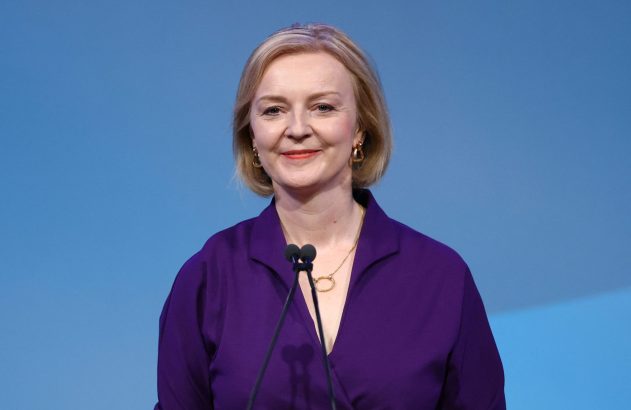 Liz Truss em roxo, sorrindo à frente de microfones com fundo azul.