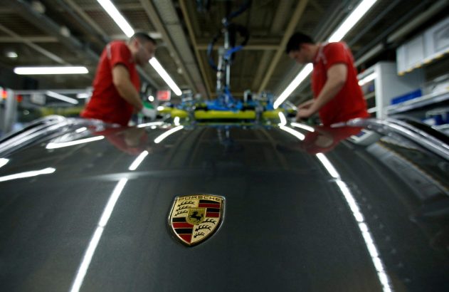 Logotipo da Porsche em carro. Trabalhadores em vermelho, refletidos, montam veículo em fábrica.