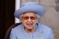 Rainha Elizabeth II de chapéu azul e óculos, sentada e sorrindo com colar de pérolas e broches.