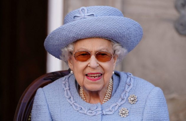 Rainha Elizabeth II de chapéu azul e óculos, sentada e sorrindo com colar de pérolas e broches.