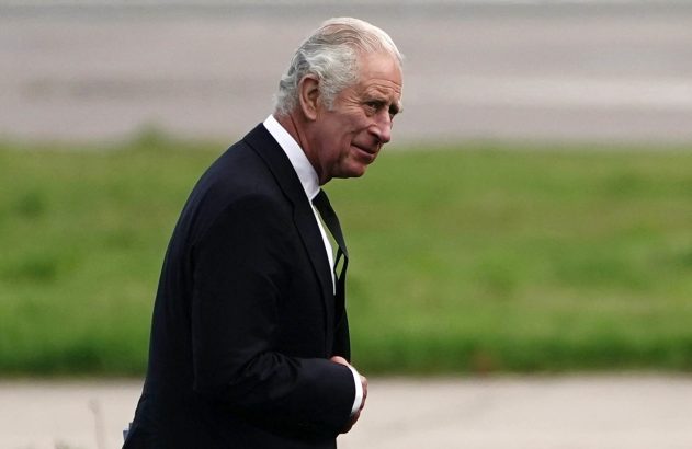 Rei Charles III, em terno preto, caminha e olha para a direita, mãos à frente. Fundo desfocado de grama e estrada.