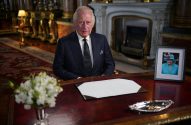 Rei Charles III sentado à mesa com foto da Rainha Elizabeth II e documento em branco. Ambiente palaciano.