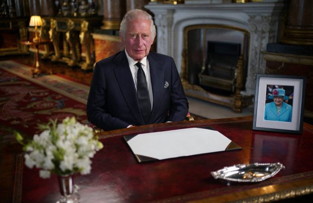 Rei Charles III sentado à mesa com foto da Rainha Elizabeth II e documento em branco. Ambiente palaciano.