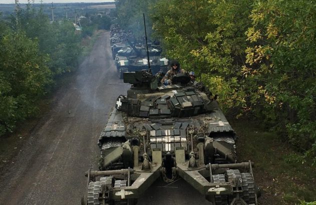 Comboio de tanques ucranianos com rolo de minas em estrada de terra arborizada.