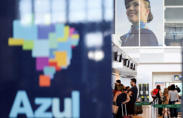Aeroporto com logo da Azul Linhas Aéreas em primeiro plano, passageiros fazem check-in e pôster de aeromoça ao fundo.