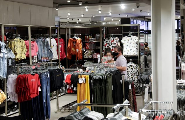 Clientes usando máscaras em loja de roupas, observando peças em cabides e prateleiras.