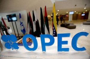 Logotipo azul da OPEC e seu símbolo, com bandeiras de países membros ao fundo em um salão.
