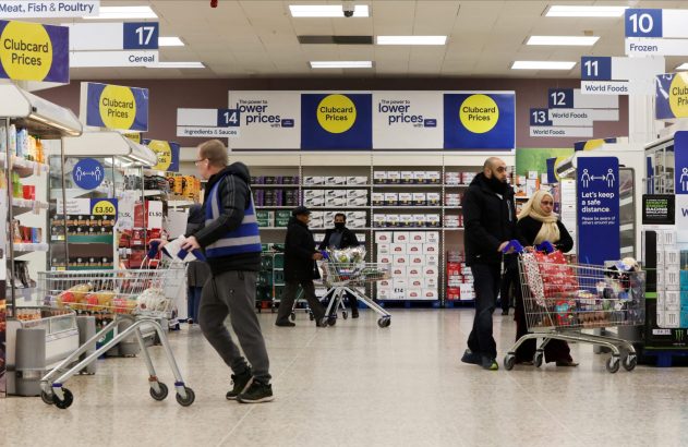 Clientes com carrinhos em corredor de supermercado Tesco, com avisos de preços promocionais e distanciamento social.