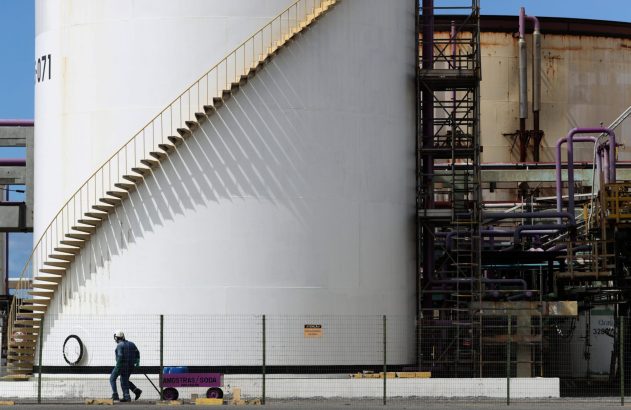 Tanque industrial branco com escada em espiral e tubulações. Um trabalhador puxa um carrinho em frente à estrutura.