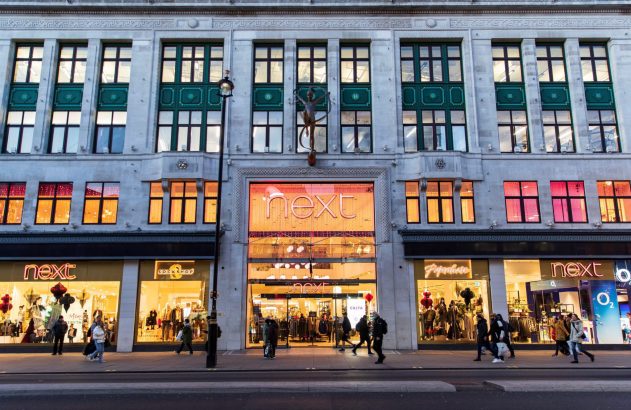 Fachada de loja Next com letreiros e vitrines iluminados, pessoas na calçada e estátua central.
