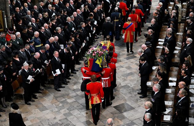 Funeral em catedral: caixão real coberto por bandeira e flores, carregado por militares entre convidados de luto.