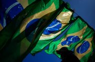 Múltiplas bandeiras do Brasil tremulam sob céu azul. Silhueta de pessoa as observa no canto inferior direito.