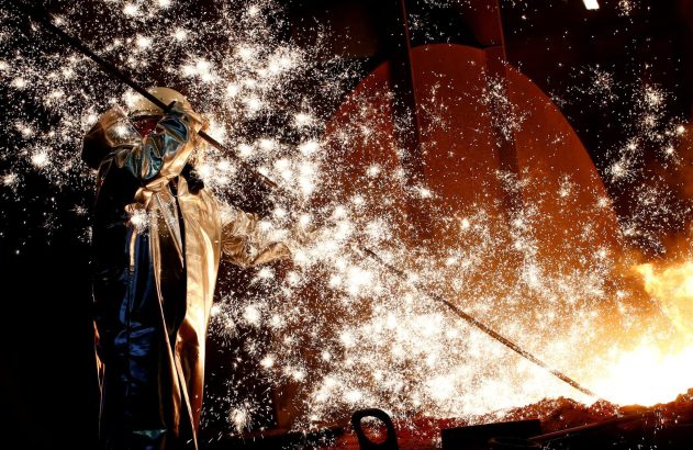 Pessoa em traje prateado manipula metal fundido, gerando intensa chuva de faíscas em fornalha industrial.