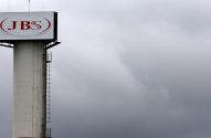 Torre de concreto branco com letreiro JBS vermelho no topo, sob céu cinza e nublado.
