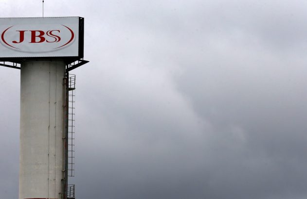 Torre de concreto branco com letreiro JBS vermelho no topo, sob céu cinza e nublado.