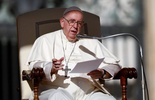 Papa Francisco, de batina branca e cruz peitoral, sentado, falando em microfone e segurando papéis.