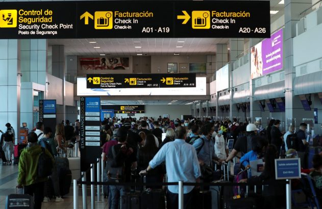 Multidão espera em fila para check-in ou segurança em aeroporto. Placas aéreas guiam os passageiros.