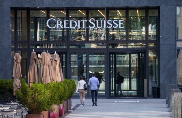 Entrada do Credit Suisse: fachada de vidro, pessoas se aproximando e guarda-sóis dobrados.