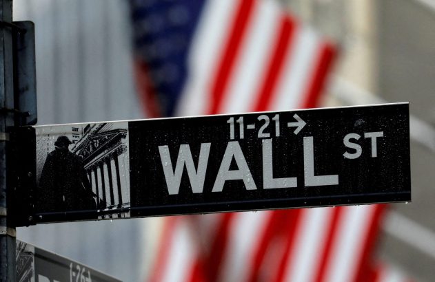 Placa de rua "Wall St" molhada, com uma bandeira dos EUA desfocada ao fundo.