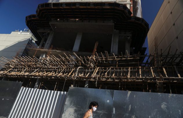 Canteiro de obras: prédio em construção com andaimes. Uma mulher de máscara caminha em frente.