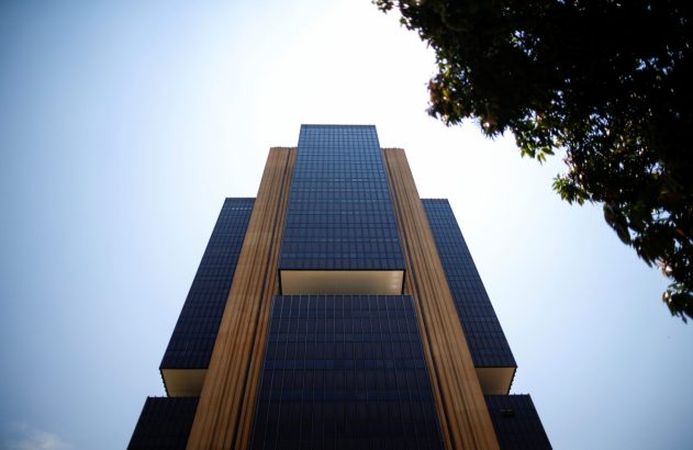 Vista de baixo de um edifício moderno com fachada de vidro escuro e colunas claras. Céu azul e folhagem à direita.