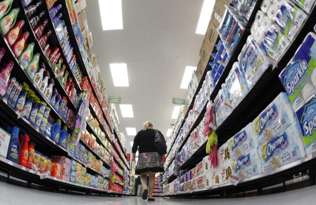 Visão de baixo: mulher em corredor de supermercado com prateleiras cheias de produtos de limpeza e higiene.