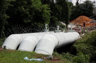 Grandes tubulações brancas deitadas na grama. Ao fundo, árvores e um monte de terra vermelha com duas pessoas.