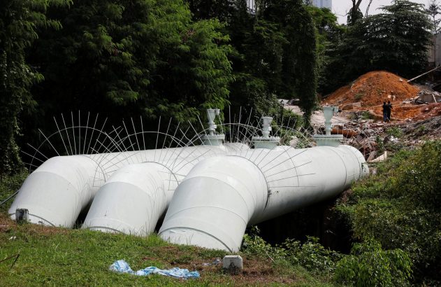 Grandes tubulações brancas deitadas na grama. Ao fundo, árvores e um monte de terra vermelha com duas pessoas.
