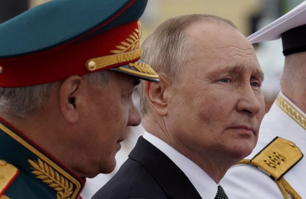 Vladimir Putin em perfil, olhando à direita, ao lado de um oficial em uniforme militar e outro militar à direita.