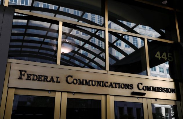 Entrada dourada da Federal Communications Commission. Letreiro e reflexos de prédio, luzes internas e número 445 na janela.