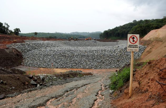 Canteiro de obras com grande montanha de rochas, estrada de terra lamacenta, máquinas e sinal 'SENTIDO PROIBIDO'.