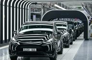 Fila de carros Tesla pretos, o primeiro com placa GIGA 001, em linha de produção de fábrica. Reflexos de luzes nas carrocerias.