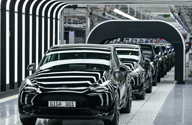 Fila de carros Tesla pretos, o primeiro com placa GIGA 001, em linha de produção de fábrica. Reflexos de luzes nas carrocerias.