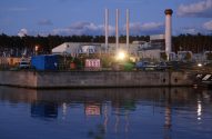 Terminal de gás natural com chaminés e tubulações brancas, refletido na água azul escura ao anoitecer.