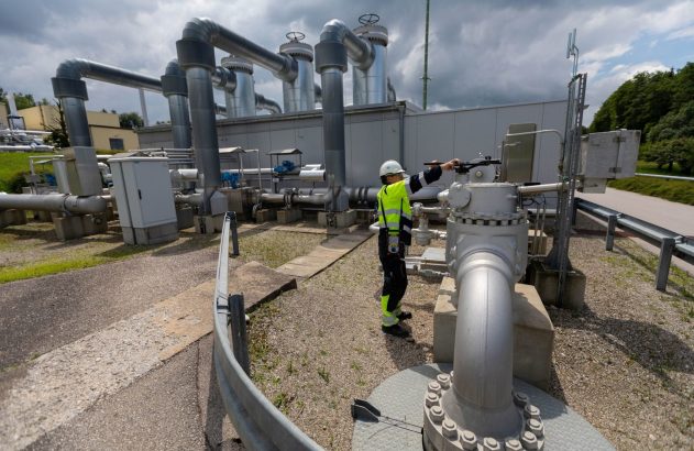 Técnico de segurança ajusta válvula de tubulação em planta industrial de gás. Grandes dutos e equipamentos ao fundo.