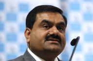Gautam Adani em um pódio com microfone, olhando para o lado. Fundo quadriculado azul claro desfocado.