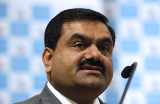 Gautam Adani em um pódio com microfone, olhando para o lado. Fundo quadriculado azul claro desfocado.