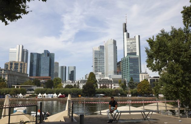 Um homem sentado à beira de um rio com skyline de arranha-céus ao fundo.