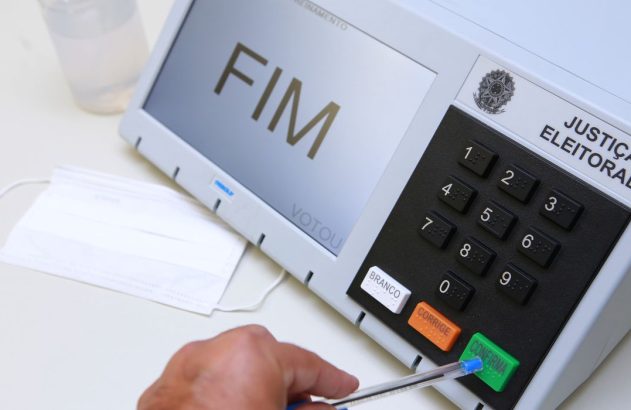 Mão confirma voto em urna eletrônica brasileira. Tela mostra 'FIM VOTOU'. Máscara e álcool gel.