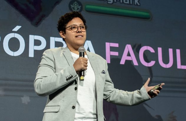 Palestrante de óculos e blazer cinza segura microfone e gesticula. Ao fundo, painel com texto "ÓP" e "FACUL".