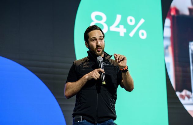 Homem em palco segurando microfone e gesticulando. Ao fundo, tela verde exibe '84%' e tela azul.