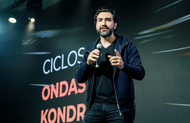 Palestrante segurando microfone e gesticulando em palco, com termos 'CICLOS' e 'ONDAS' projetados ao fundo.