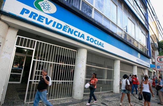 Fachada do prédio da Previdência Social com pessoas circulando na calçada em frente aos portões.